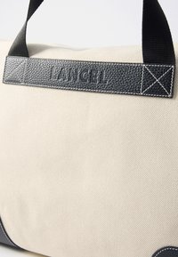 Sac bej din pânză cu curea din piele texturată neagră, având cuvântul "LANCEL" embosat. Include Cusături și accente albe contrastante.