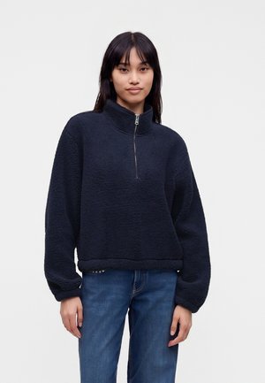 Jeune femme aux longs cheveux foncés portant un pull polaire bleu marine avec une fermeture éclair à l’avant et un jean bleu sur un fond clair uni.