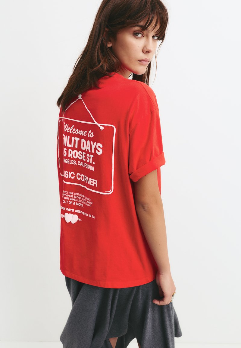 Tricou roșu supradimensionat cu mâneci rulată, având un text grafic alb pe spate, purtat peste o fustă gri închis. Țesătură din bumbac, croi lejer.