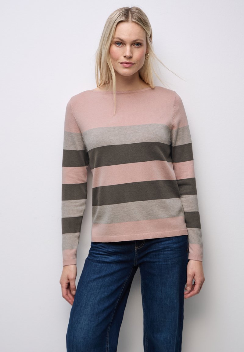 Femme aux longs cheveux blonds portant un pull rayé rose, gris et foncé ainsi qu'un jean bleu, debout contre un mur blanc.