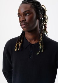 Polo shirt negru cu mânecă lungă și guler, având patru butoane și o textură moale. Remarcabil pentru accentele de păr împletit colorat.