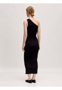 Zwarte geribbelde jurk met een één-schouder ontwerp, aansluitende silhouet en een zoom die de enkels raakt. Gekleed met zwarte platte schoenen.