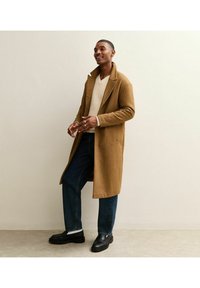 Brauner Wollmantel mit Reverskragen, getragen über einem cremefarbenen V-Ausschnitt-Pullover. Dunkelblaue Jeans mit lockerer Passform und schwarze Slipper.