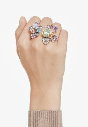 Guldtonede ringe formet som blomster, med multikolorede ædelsten og kompleks pavé-detaljering. Hånd iført ringe med en lys hudfarve.