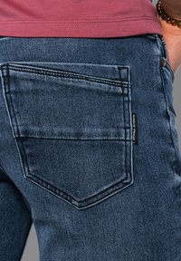 Jeans de mezclilla azul oscuro con un diseño distintivo en el bolsillo trasero. Presentan costuras visibles y una pequeña etiqueta cosida en la costura lateral.