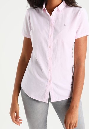 Frau trägt ein hellrosa kurzärmeliges Hemd mit Knöpfen und hellgraue Hose, steht mit entspannt an den Seiten hängenden Armen.