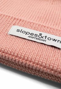 Różowa dzianinowa czapka beanie o prążkowanej teksturze, z białą metką z napisem "slopes&town AMSTERDAM" w czarnym kolorze.