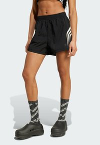 Fekete sportos rövidnadrág, texturált felülettel, oldalain három fehér csíkkal. Fekete slip-on klumpákkal és mintás zoknikkal párosítva.