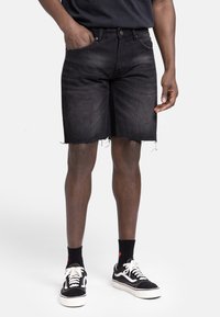 Shorts en denim noir avec un ourlet effiloché, cinq poches et fermeture à bouton métallique. Porté avec des baskets noires et des chaussettes sombres.