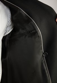 Interno di giacca nera con una texture liscia, tasca con zip e piping chiaro a contrasto. Camicia a righe visibile accanto.