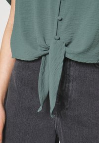 Haut vert en tissu léger avec un détail noué à l'avant et des boutons le long de la patte de boutonnage, associé à un jean en denim foncé.