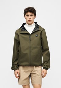 Άνδρας φορώντας ένα πράσινο χακί μπουφάν με φερμουάρ πάνω από ένα λευκό ζιβάγκο, σε συνδυασμό με μπεζ σορτς μέχρι το γόνατο, στέκεται μπροστά σε ένα απλό φόντο.