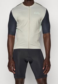 Camisa de ciclismo gris claro con mangas cortas en marino y cremallera, combinada con pantalones cortos negros que tienen una zona acolchada. Tela lisa y elástica.