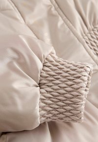 Chaqueta acolchada en suave satén beige, con detalle de puños fruncidos. La tela tiene una textura suave con un brillo sutil.