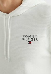 Felpa bianca a coste con cappuccio e cordini regolabili. Presenta il logo "TOMMY HILFIGER" con dettagli neri e rossi sul petto. Tessuto morbido.