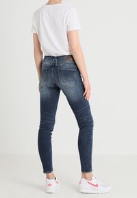 Jeans de mezclilla azul ajustados con cintura alta, diseño de cinco bolsillos y un ligero desvanecimiento. Combinados con una camiseta blanca y zapatillas con detalles en rosa.