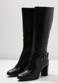 Mustat nahkaiset polvipituiset saappaat, joissa on lohkojäykät korkokengät ja sivuvedin, ja jotka on varustettu koristeellisella soljella nilkan kohdalla.