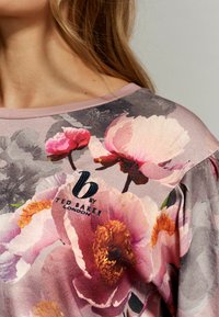 Bloemenpatroon top in een zachte stof, met roze en grijze tinten, voorzien van een geborduurd logo in donkerblauw op het borstgedeelte.