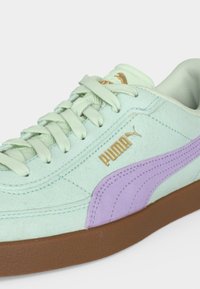 Ανοιχτό πράσινο αθλητικό παπούτσι Puma με λωρίδα λαβάντας στο πλάι, χρυσό λογότυπο Puma, άσπρα κορδόνια και καφέ λαστιχένια σόλα σε κοντινή λήψη.