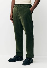 Green corduroy trousers with a straight fit, featuring a button closure and side pockets, paired with black lace-up shoes.