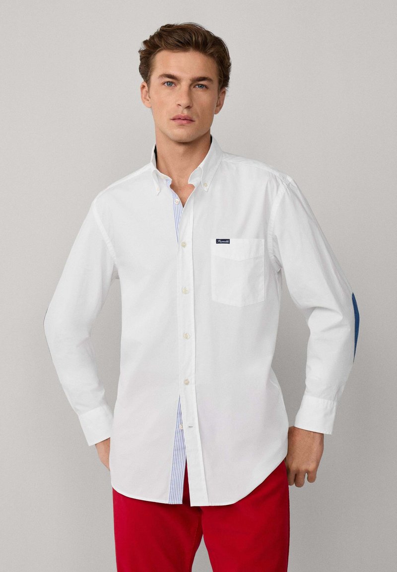 Chemise blanche à manches longues avec col boutonné, présentant une patte de boutonnage rayée, une poche et un logo sur la poitrine. Pantalon rouge.