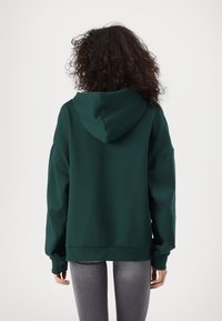 Sudadera verde con capucha en un corte relajado, que presenta un bolsillo tipo canguro y puños acanalados. La tela es suave con una textura lisa.