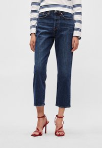 Τζιν παντελόνι μπλε από denim με ψηλή μέση και άνετη εφαρμογή, με κομμένο μήκος στο πόδι, σε συνδυασμό με κόκκινες σανδάλες με λουράκια και ψηλό τακούνι.