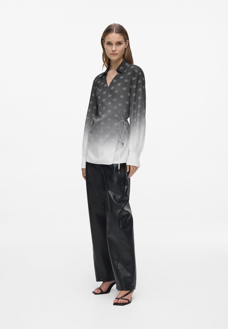 Jeune femme portant un chemisier portefeuille dégradé noir et blanc, un pantalon en cuir noir à jambes larges et des sandales noires à talons avec des lanières, debout devant un fond blanc.