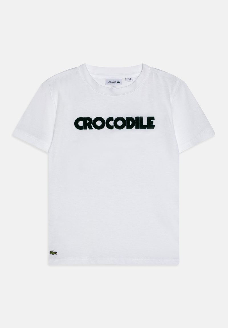 T-shirt en coton blanc avec "CROCODILE" en texte noir en gras, manches courtes, et un petit logo Lacoste brodé à l'ourlet.