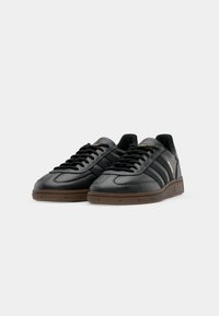 Sneakers in pelle nera con tre strisce scure, suola in gomma marrone, design con lacci e dettagli del logo dorato di lato.
