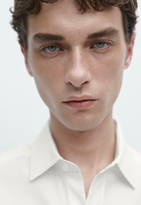 Primo piano di una persona che indossa una camicia bianca con colletto. La camicia presenta un design con bottoni, una texture liscia e uno sfondo neutro che enfatizza il volto.