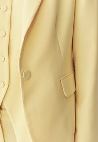 Gros plan sur un blazer beige sur mesure avec un bouton assorti et un détail de poche à rabat sur la manche et à l'avant.