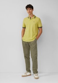 Geel poloshirt met donkere gestreepte kraag, gecombineerd met olijfgroene corduroypantalon en casual witte sneakers met houten zolen.