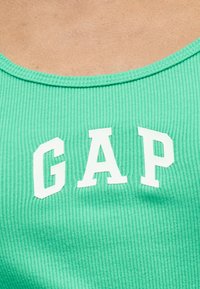 Κοντινό πλάνο ενός ανοιχτό πράσινου ριγωτού τοπ χωρίς μανίκια με τολμηρά λευκά γράμματα "GAP" στο κέντρο του υφάσματος.