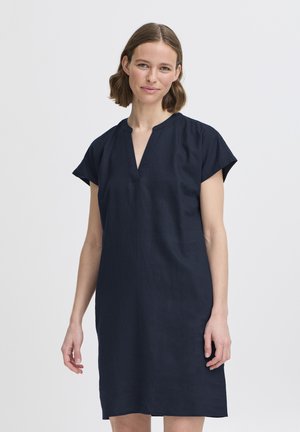 Žena s krátkymi hnedými vlasmi v uvoľnených tmavomodrých krátkych šatách s výstrihom do V stojaca pred bielym pozadím.