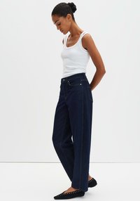 Jeans blu scuro a vita alta con gambe dritte, abbinati a una canottiera bianca aderente. Scarpe basse nere con borchie decorative.