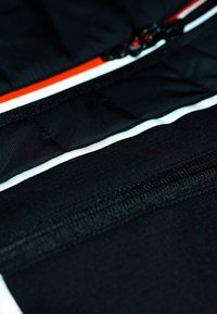 Schwarzer gesteppter Stoff mit hervorgehobenen, markanten horizontalen weißen und orangefarbenen Streifen. Verfügt über eine zentrale Reißverschluss-Detail und eine glatte Textur.