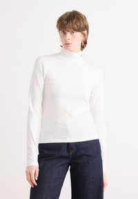 White long-sleeve turtleneck top in a fitted design, made of smooth fabric with a simple neckline and minimal detailing, paired with dark jeans.