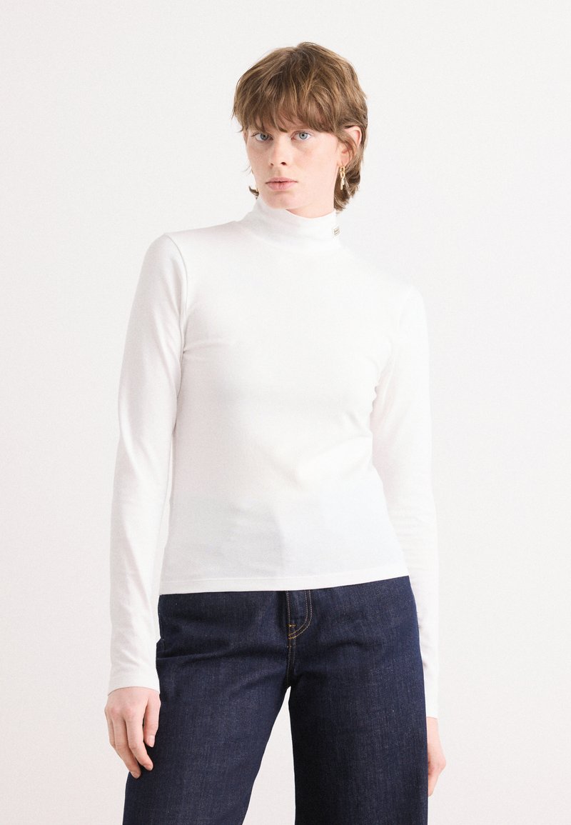 White long-sleeve turtleneck top in a fitted design, made of smooth fabric with a simple neckline and minimal detailing, paired with dark jeans.