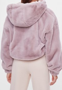 Persoon in een cropped, lila fuzzy hoodie met lange mouwen en beige broek, gezien vanuit de rug.