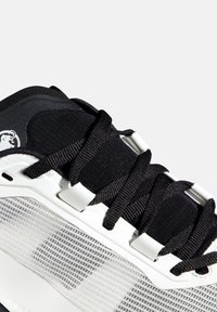 Κοντινό πλάνο μαύρου και άσπρου αθλητικού παπουτσιού με μαύρα κορδόνια, πλευρικά πάνελ από δίχτυ και επενδεδυμένη γλώσσα, σε λευκό φόντο.