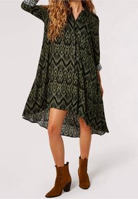 Grünes gemustertes Hemdkleid mit durchgehender Knopfleiste, langen Ärmeln und asymmetrischem Saum. Getragen mit braunen Stiefeletten. Strukturierten Stoff.