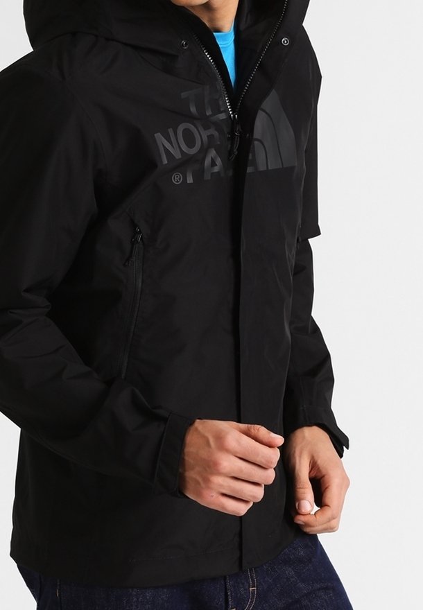 Schwarze wasserdichte Jacke mit Kapuze, Reißverschluss und einem großen Logo auf der Brust. Verfügt über seitliche Reißverschlusstaschen und eine glatte Textur.