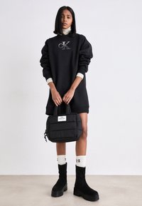 Mujer con vestido sudadera negro de Calvin Klein, cuello alto blanco, botas negras con plataforma, calcetas blancas con el número 23, sosteniendo un bolso acolchado negro.