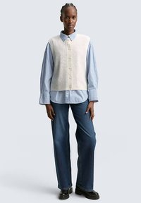 Krémový pletený sveter bez rukávov cez modro-bielo pruhovanú košeľu na gombíky, doplnený širokými modrými džínsami a čiernymi čižmami.