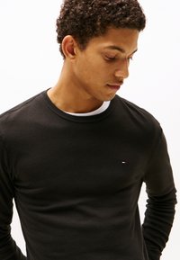 Junger Mann mit lockigem Haar trägt ein schwarzes Langarmshirt über einem weißen Unterhemd und schaut nach unten links vor weißem Hintergrund.