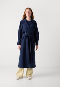 Manteau trench bleu marine avec un design à double boutonnage, taille ceinturée, grands revers et manches longues, associé à un pantalon large jaune clair.