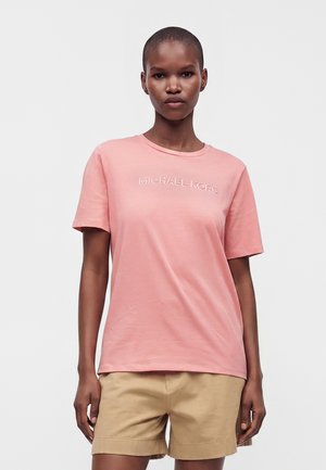 Kobieta ubrana w różowy T-shirt Michael Kors i beżowe szorty, stojąca na jednolitym białym tle.