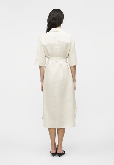 Γυναίκα με μακριά σκούρα μαλλιά που φοράει μπεζ μεσήλικο φόρεμα με μανίκια μέχρι τον αγκώνα, ζώνη και μαύρα πέδιλα με τακούνι, εμφανιζόμενη από πίσω.