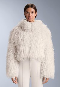 Biały, futrzany krótki płaszcz z wysokim kołnierzem i długimi rękawami. Teksturowany materiał nadaje puszysty wygląd, zestawiony z dopasowanymi jasnymi spodniami.